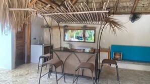 Une pièce rustique avec un toit de chaume, un petit coin bar avec des tabourets, un évier et une fenêtre donnant sur la verdure.
