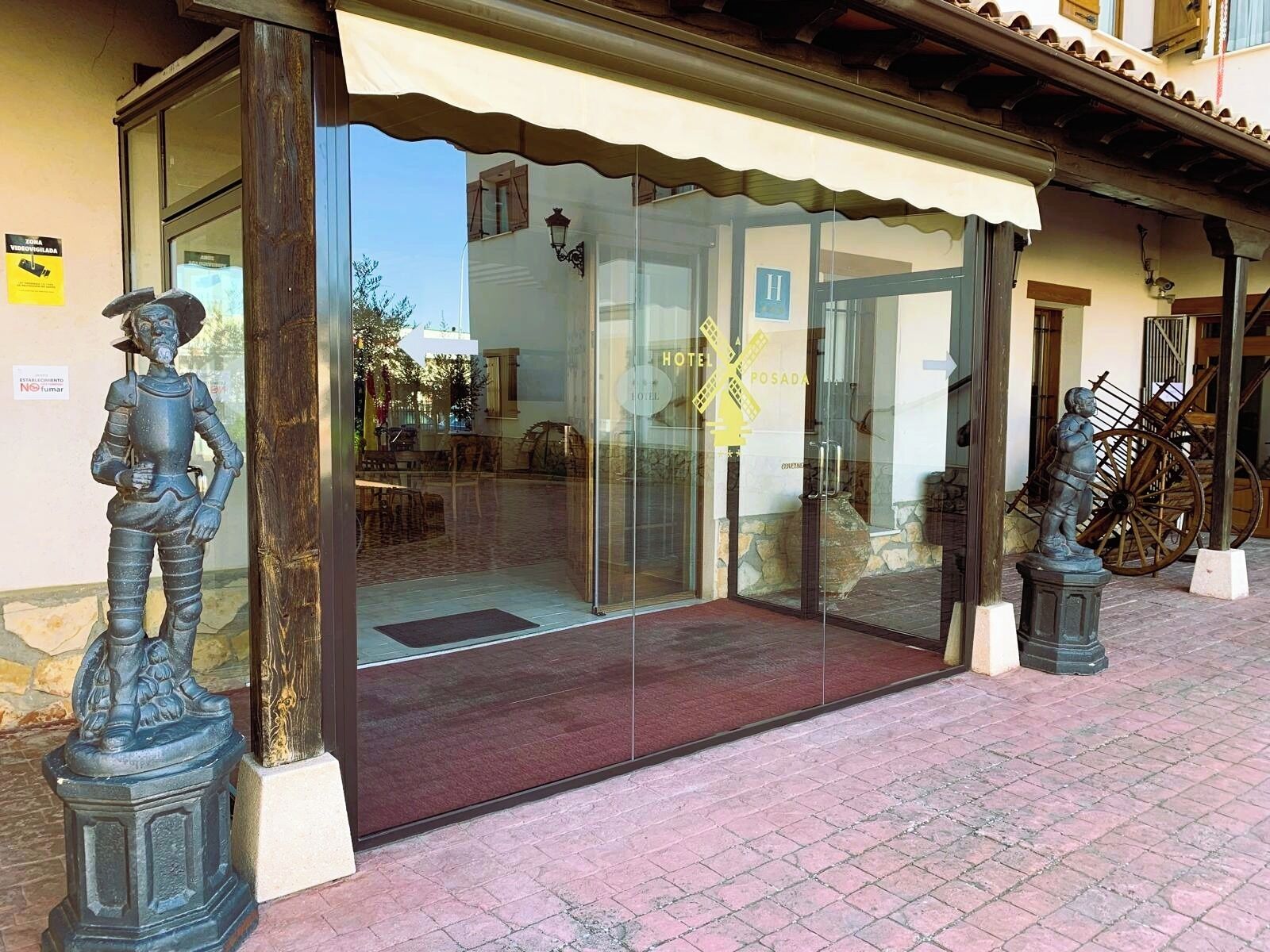 Entrada de un hotel con puerta de cristal, una estatua de piedra a la izquierda y un carrito de madera a la derecha.