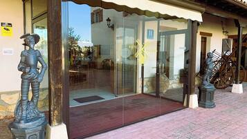 Entrada de un hotel con puerta de cristal, una estatua de piedra a la izquierda y un carrito de madera a la derecha.