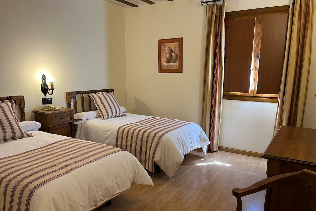 A bedroom with two beds, wooden furniture, a nightstand with a lamp, and a window with curtains.