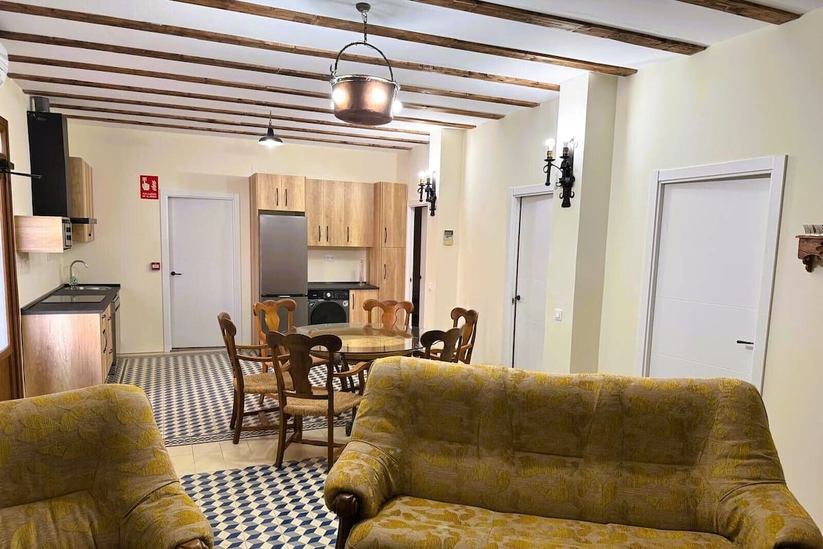 A living room with a checkered floor, a sofa, a dining table with chairs, a kitchen with wooden cabinets, and a hanging light fixture.