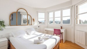 A bedroom with a bed, a desk, and a chair. There is a window with a view of buildings and a plant on the left side.