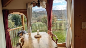 Una mesa de comedor de madera con sillas en una habitación con vigas de madera en el techo, ventanas amplias con cortinas y vistas a un paisaje verde.