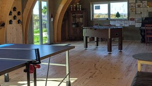 Una habitación amplia con mesa de ping pong, mesa de billar y piano. La habitación tiene techos de madera y ventanas grandes.