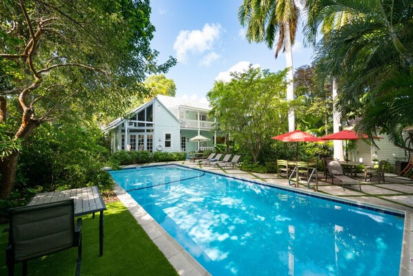 A swimming pool with lounge chairs and umbrellas, surrounded by lush greenery and a white building with a balcony. - Family Reunion Compound - Great for Groups! Large pool. (Key West)
