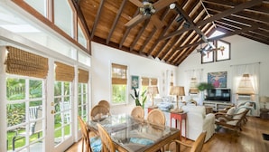A spacious living area with a glass dining table, wicker chairs, a television, and a large window overlooking a garden. - Family Reunion Compound - Great for Groups! Large pool. (Key West)