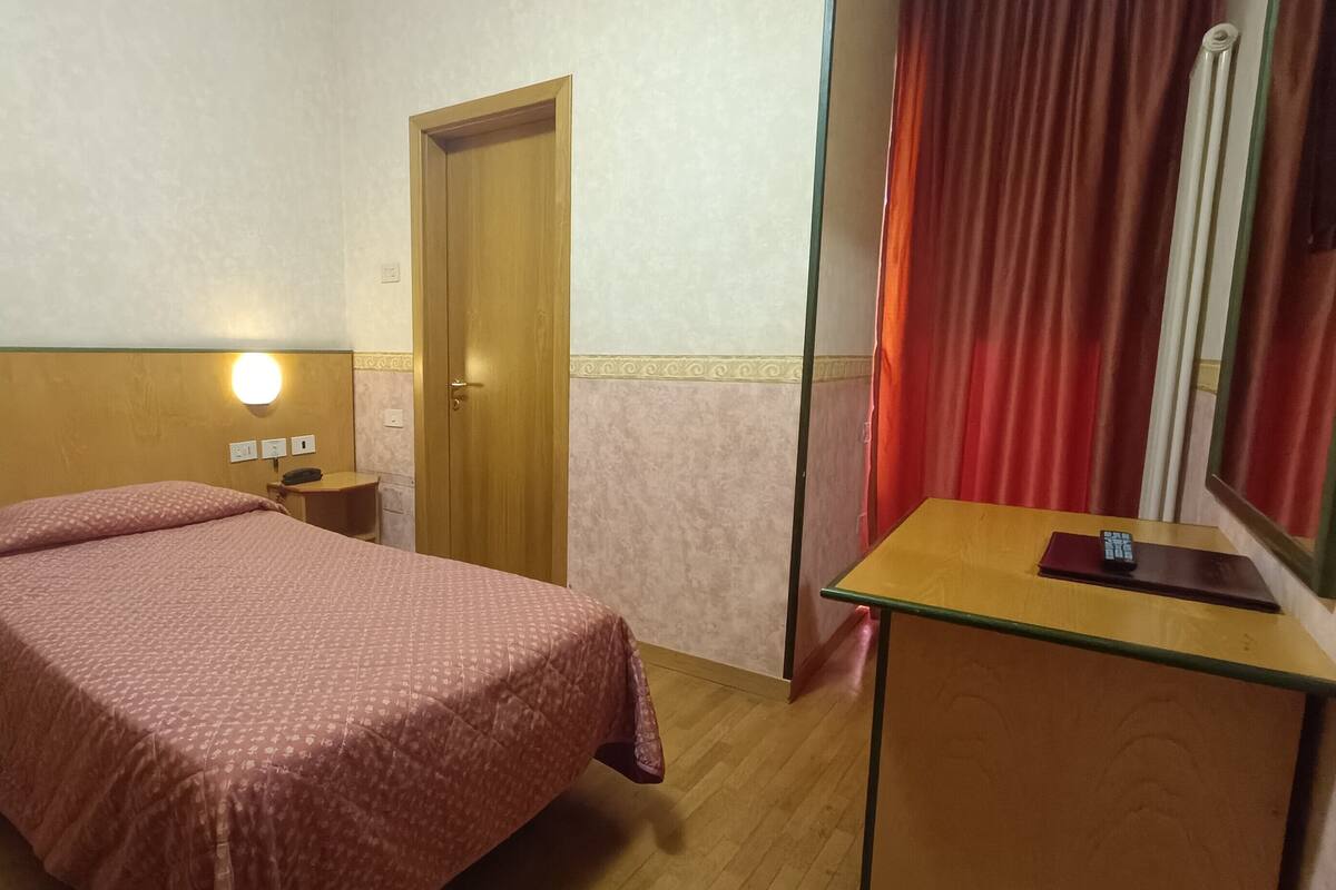 A hotel room with a bed, bedside table, and a desk with a mirror.