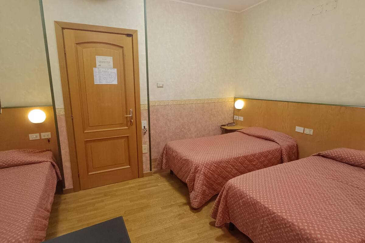 A hotel room with two single beds, a wooden door, a small bedside table with a lamp, and a wooden floor.
