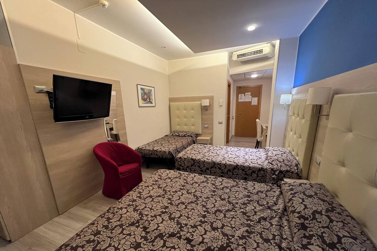 A hotel room with two beds, a red chair, a TV, and an air conditioning unit.