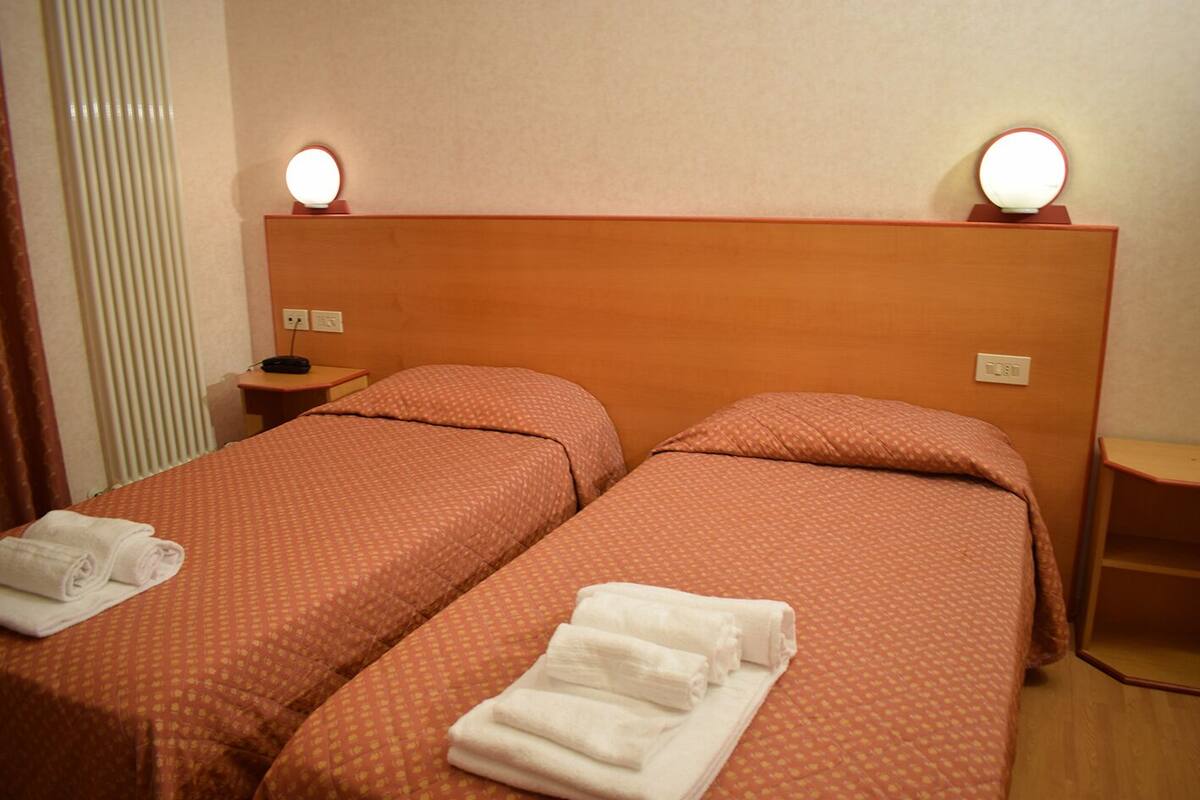 A hotel room with two beds, each with a headboard and bedside lamps. There are folded towels on the beds.