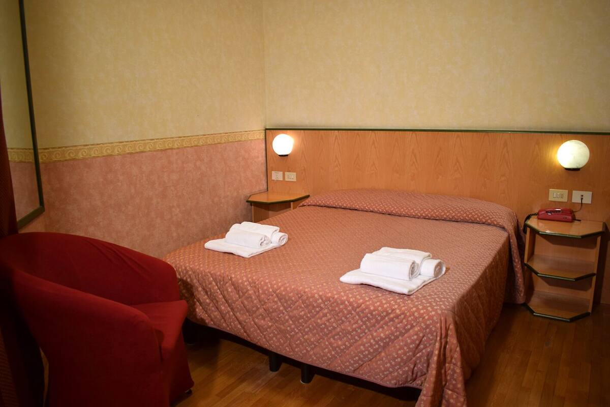 A hotel room with a bed, two bedside tables, a red armchair, and a nightstand with a lamp.