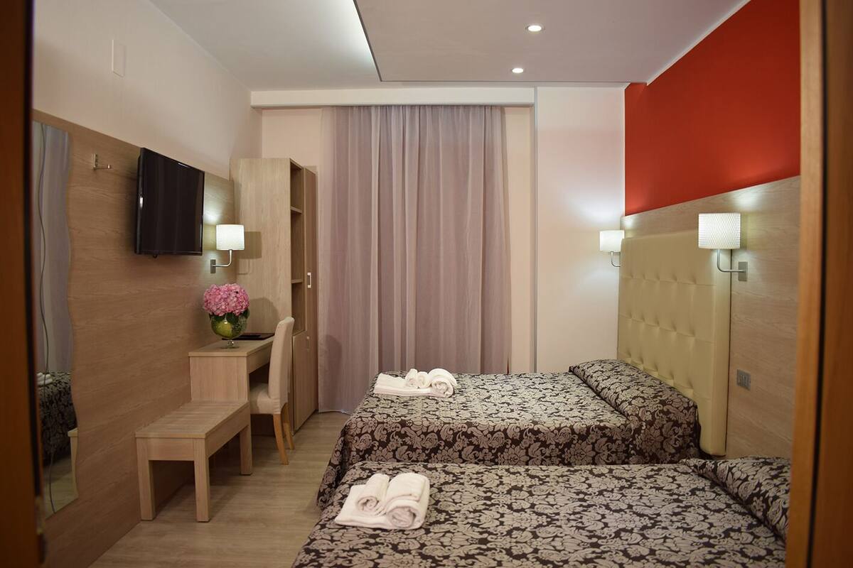 A hotel room with two beds, a desk, a chair, a TV, and a window with curtains.