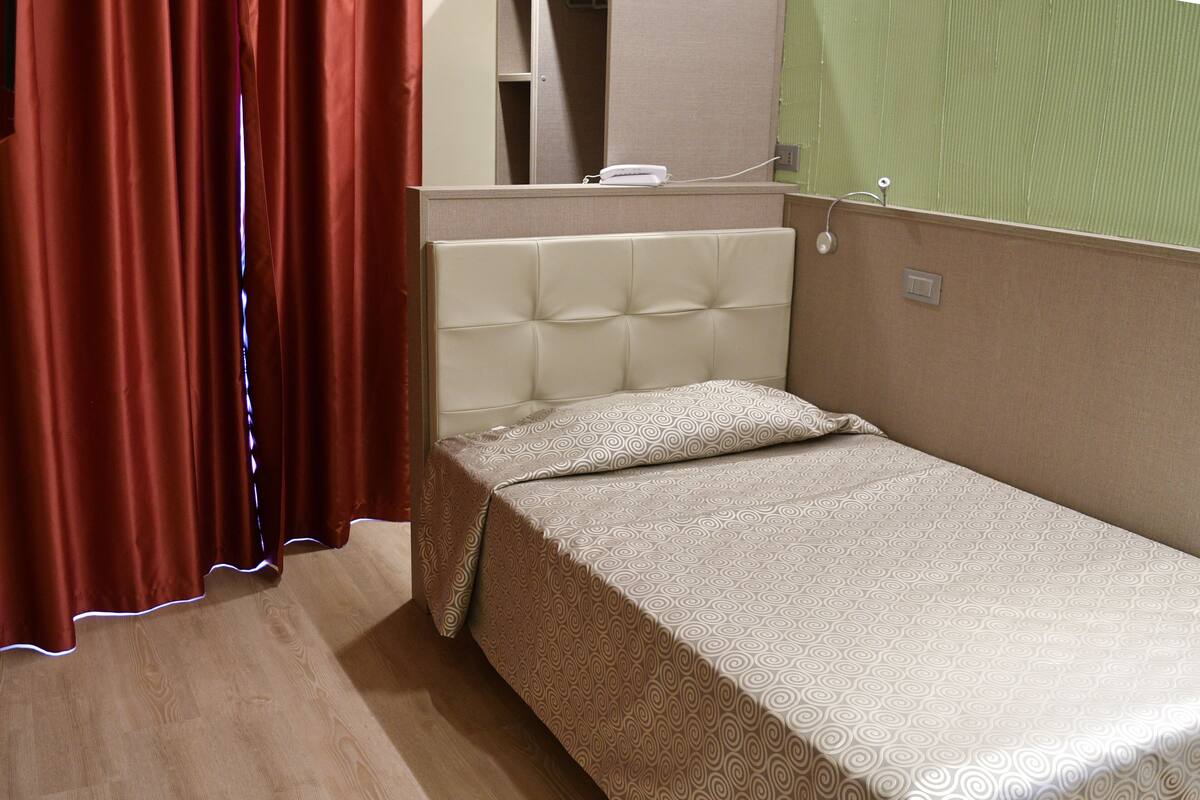 A single bed with a patterned bedspread, a tufted headboard, and a red curtain.