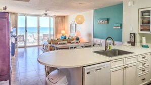 A kitchen with a central island, a sink, and a dishwasher. There is a dining area with a table and chairs, and a view of the ocean through large windows.