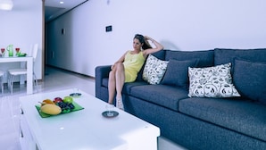 A modern living room with a grey sofa, a white coffee table with fruit, and a dining area in the background.