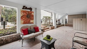 Een overdekt terras met een beige bank, een klein tafeltje met een groene decoratie en een groot bloemen schilderij aan de muur.