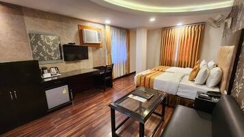 Habitación de hotel con piso de madera, cama con ropa de cama blanca y beige, escritorio con televisor de pantalla plana, cafetera y un refrigerador pequeño.