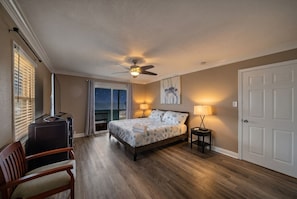 A bedroom with a bed, two bedside lamps, a ceiling fan, a desk with a chair, and a view of the outdoors.