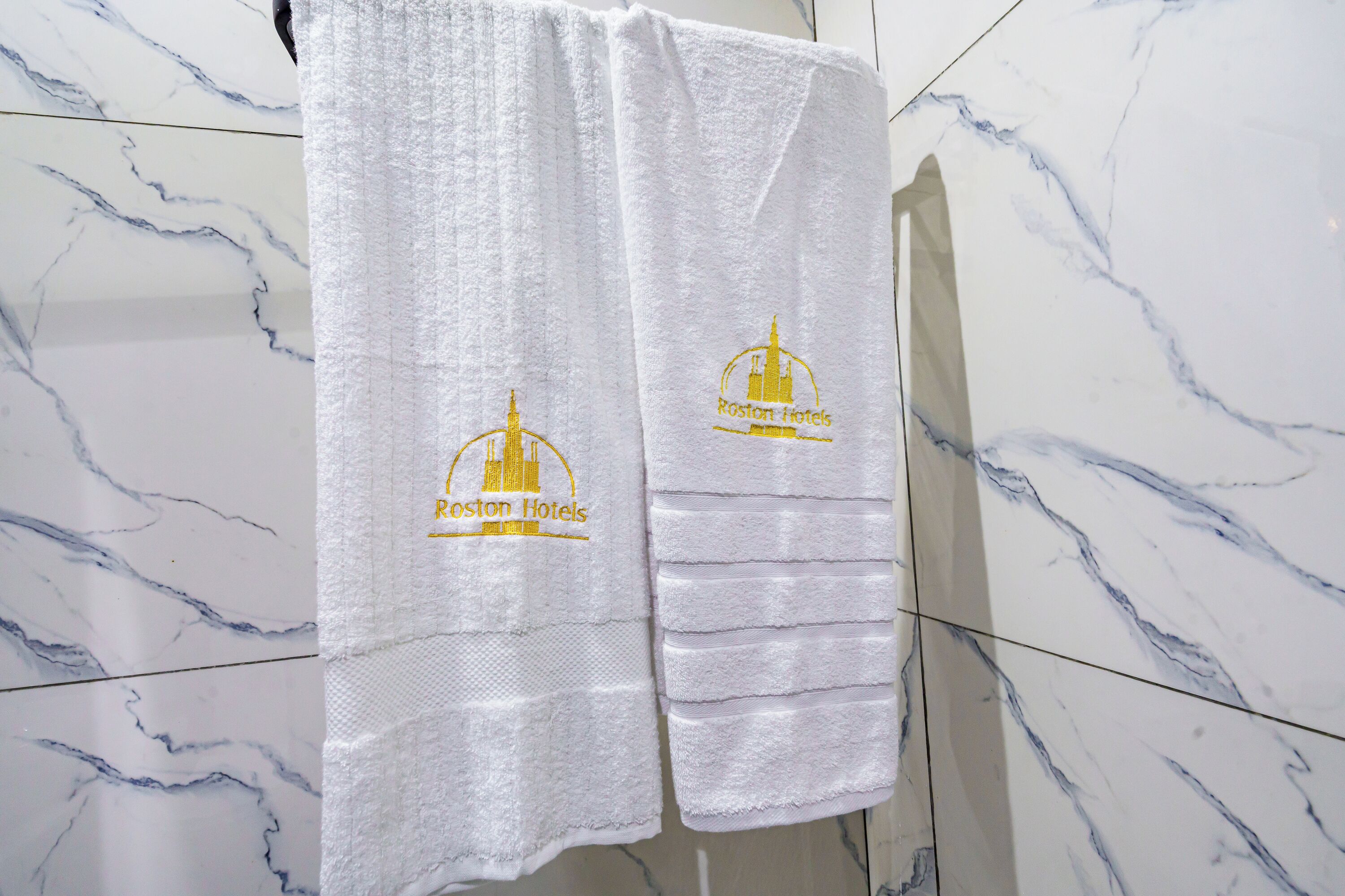 A white towel with embroidered logos of Boston Hotels hanging on a bathroom wall with marble tiles.