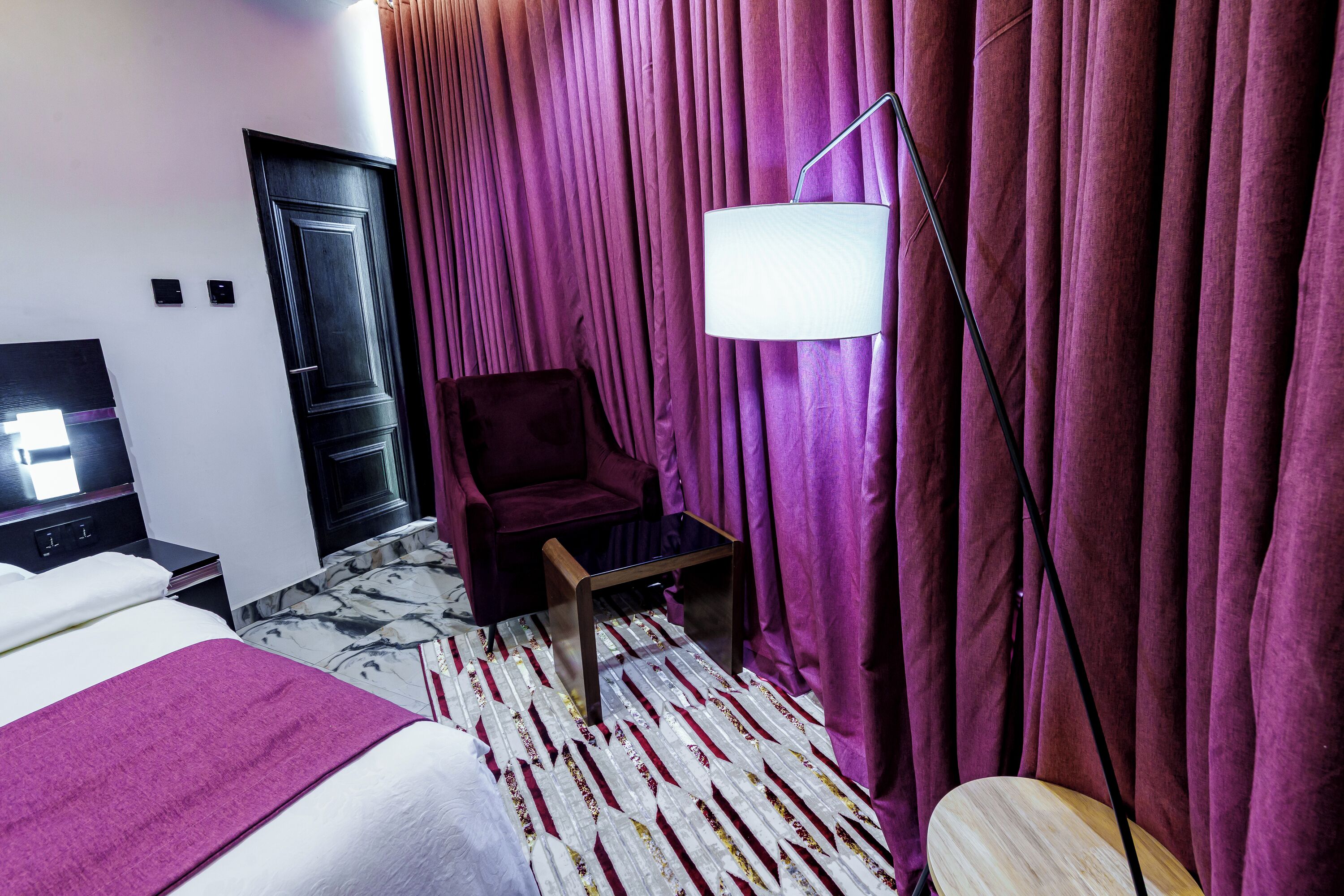 A hotel room with a bed, a chair, a small table, a lamp, and a curtain.