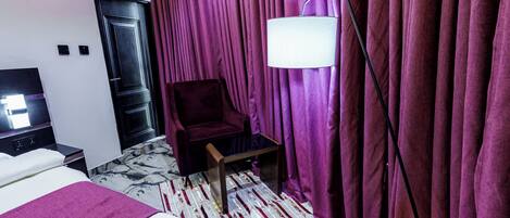 A hotel room with a bed, a chair, a small table, a lamp, and a curtain.