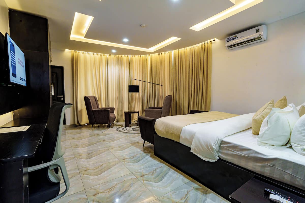 A hotel room with a bed, desk, chairs, a TV, and an air conditioning unit.