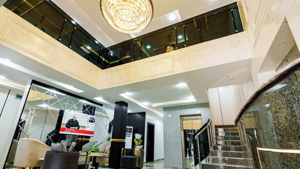 A modern lobby with a high ceiling, a chandelier, a staircase, and a reception desk. - Roston Hotels  (Port Harcourt)