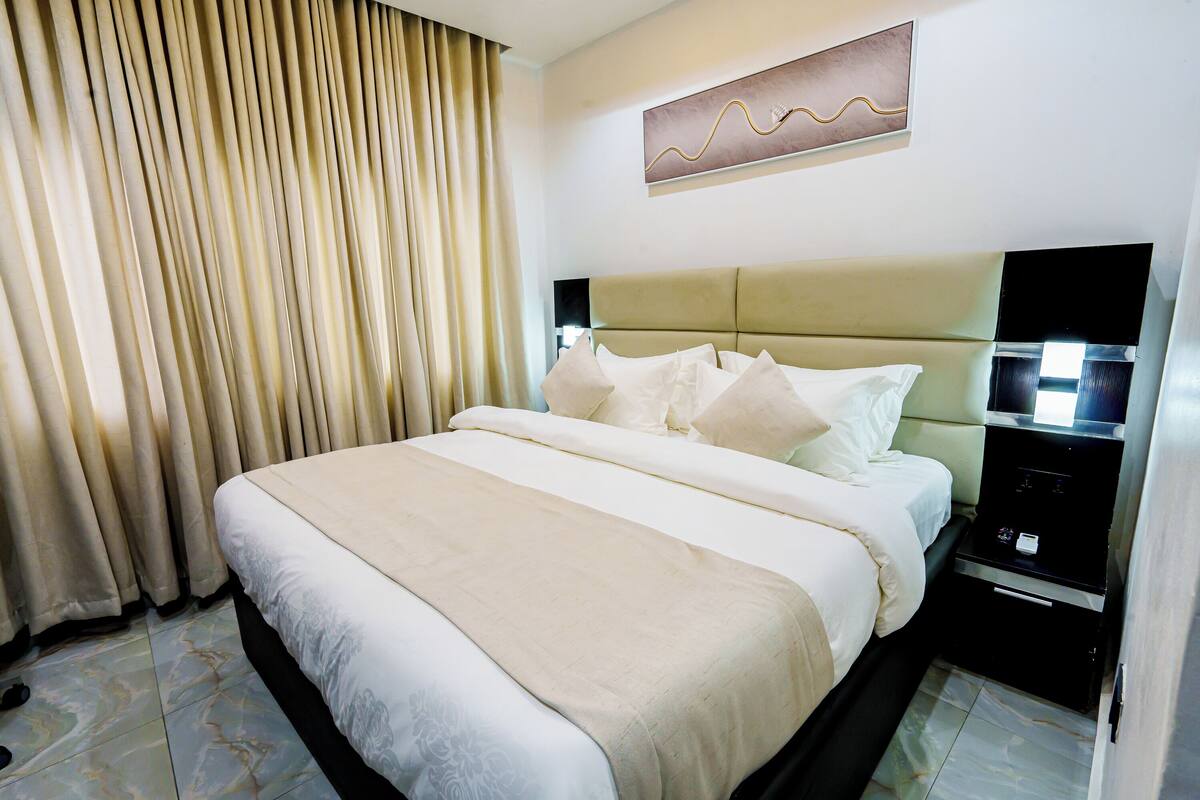 A hotel room with a large bed, a nightstand, a wall-mounted TV, and a window with curtains.