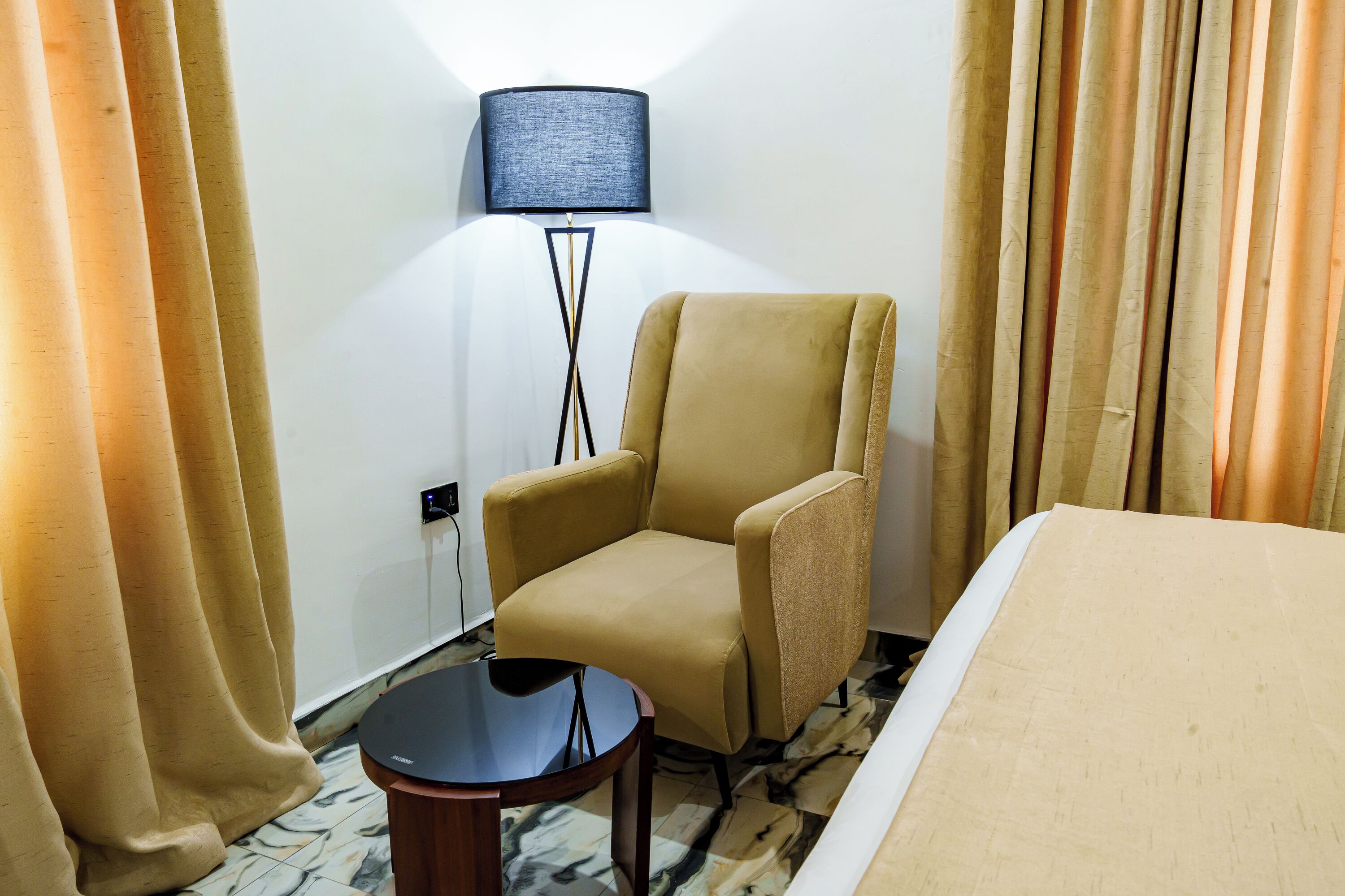 Ein Hotelzimmer mit Bett, Sessel, kleinem Tisch, Lampe und Vorhängen.