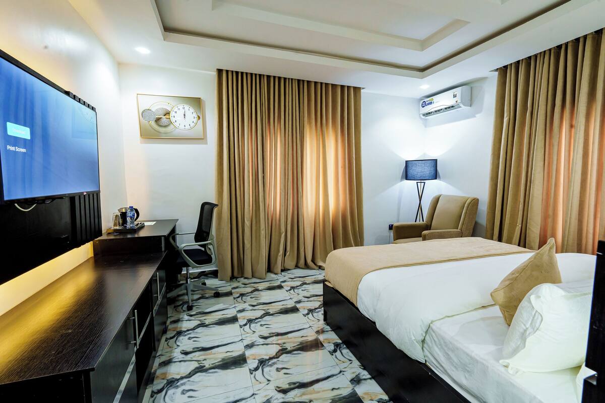 A hotel room with a bed, a desk, a chair, a television, and an air conditioning unit.