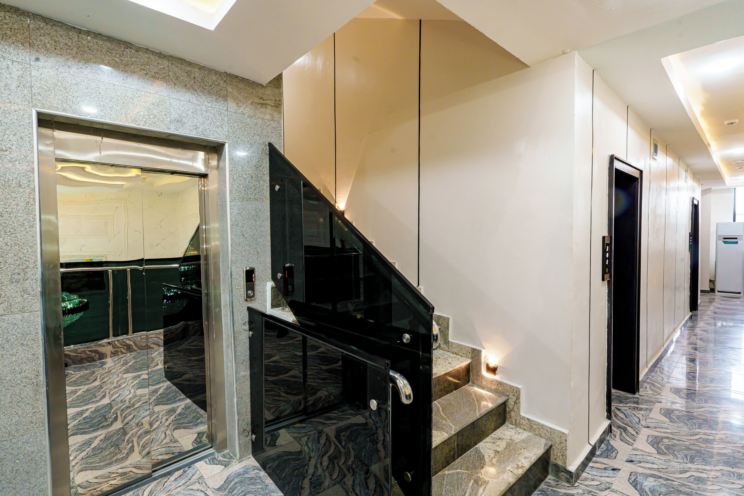 Un ascenseur moderne doté d’une porte en verre, d’un escalier en marbre et d’un couloir éclairé par un luminaire.