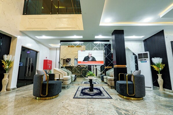 A lobby with a large screen displaying a news broadcast, seating arrangements, and a fire extinguisher. - Roston Hotels (Port Harcourt)