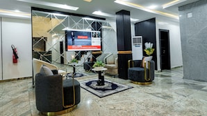 A modern lobby with a large TV screen displaying an advertisement, comfortable seating areas, and a fire extinguisher on the wall. - Roston Hotels  (Port Harcourt)