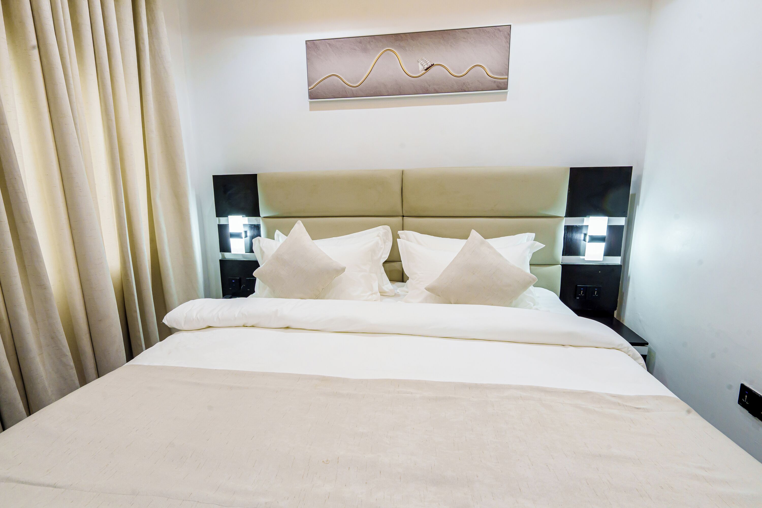 A hotel room with a large bed, a headboard with a light-colored panel, and a wall-mounted artwork above the bed.