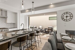 A modern kitchen with a central island, bar stools, and a dining area with a table set for four. - Olympic Escapes Gold- Desert Color Lagoon + Beaches (St. George)