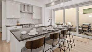 A modern kitchen with white cabinets, a central island with bar stools, and a large sliding glass door leading to an outdoor area. - Olympic Escape Silver: Desert Color Lagoon + Beaches (St. George)