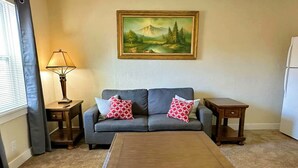 Un salon comprenant un canapé, deux tables d’appoint, une table basse, un tableau accroché au mur et une fenêtre avec des rideaux.
