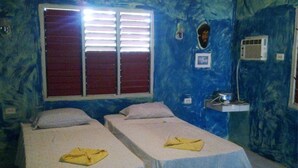 Una habitación con dos camas, un ventanal con persianas, un espejo y una pequeña estantería con una lámpara.