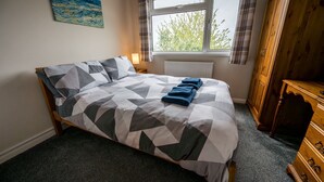 A neatly made bed with a geometric patterned bedspread, a window with a view of greenery, and a wooden wardrobe.