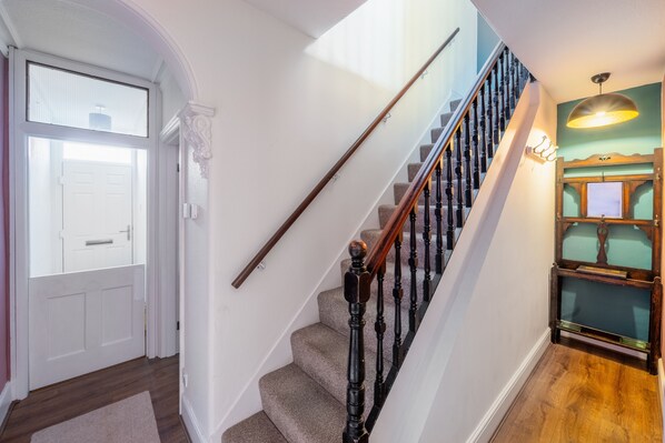Una escalera con balaustres de madera y barandilla, que lleva a un rellano con una puerta y un aplique de pared.