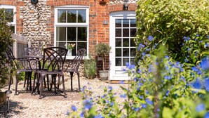 A stone building with a brick chimney, two windows, and a door. There are outdoor chairs and a potted plant near the entrance. - A delightful mid-terrace Victorian cottage only a short drive from the coast! (Docking)