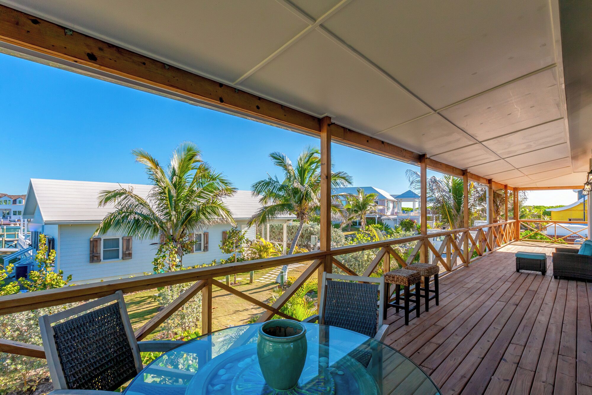 A covered outdoor area with a glass table and chairs, overlooking a residential area with palm trees and houses.