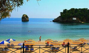 Ein Strand mit Sonnenschirmen und Liegestühlen, ein Felsen und klares, blaues Wasser.