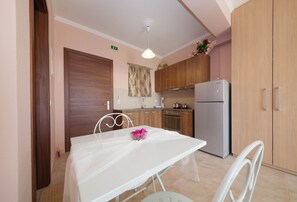 A compact kitchen with wooden cabinets, a white refrigerator, and a dining table with a white chair. - Ionian View Gem - Parga Charming Nest (Agia Kiriaki)