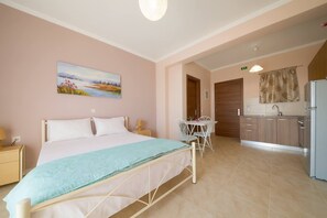 A bedroom with a bed, bedside tables, a painting, a dining area with a table and chairs, and a kitchenette with a refrigerator and cabinets. - Ionian View Gem - Parga Charming Nest (Agia Kiriaki)