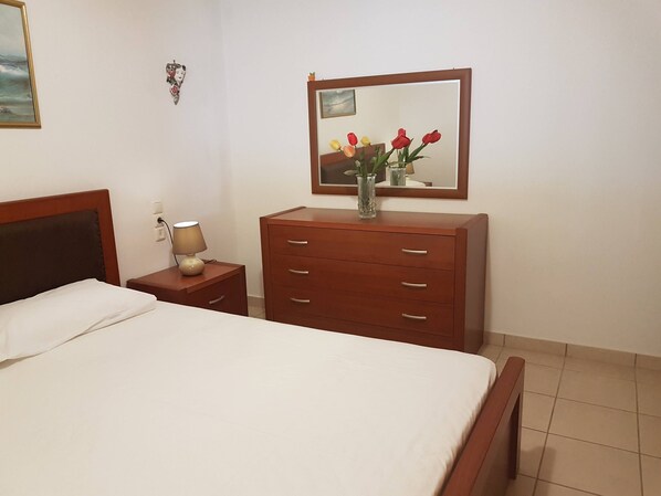 A hotel room with a bed, a wooden dresser, a nightstand with a lamp, a mirror, and a wall-mounted picture.