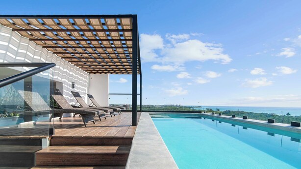 A modern outdoor swimming pool with wooden steps leading down to the water, a pergola providing shade, and a view of the ocean.