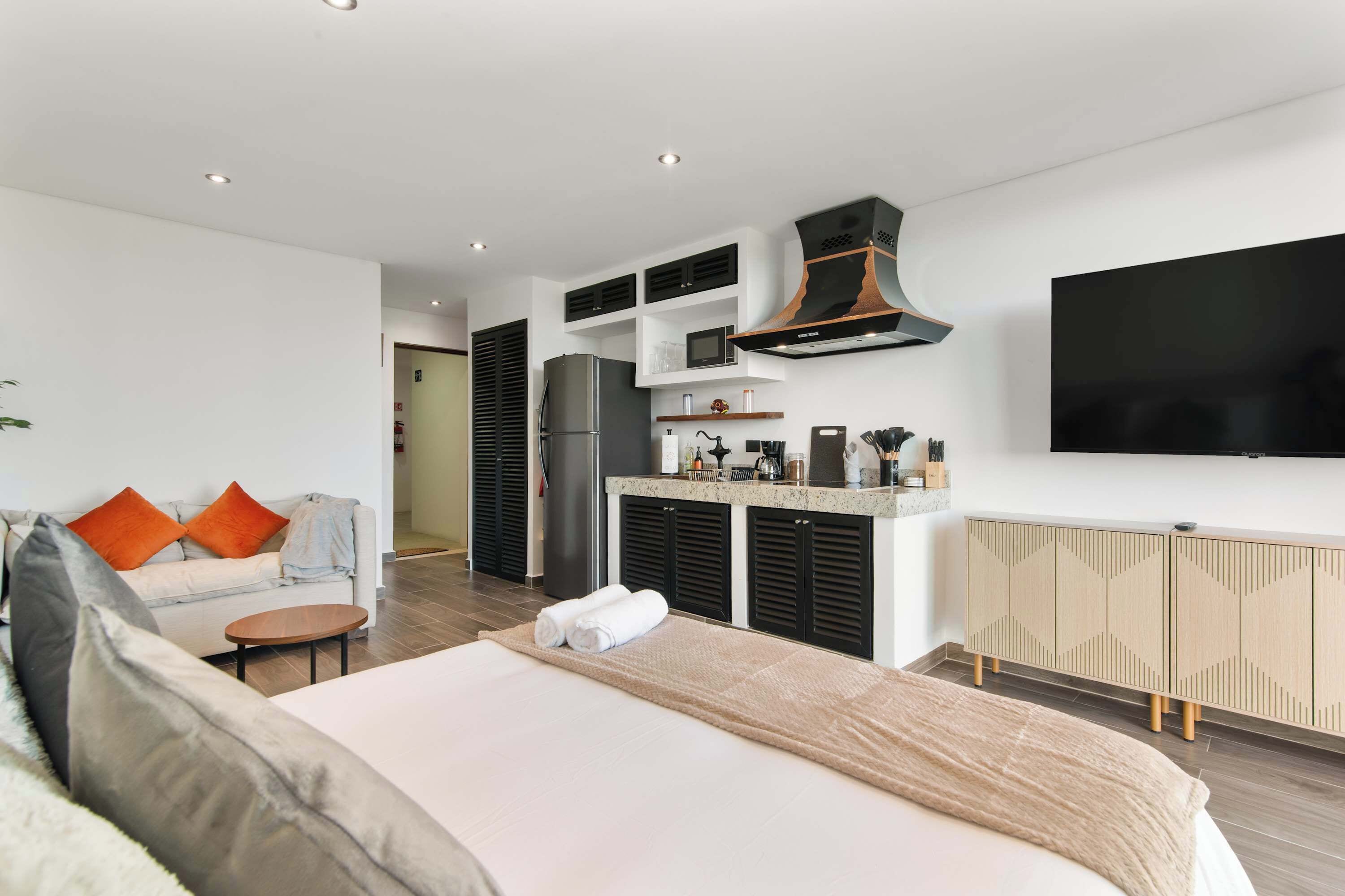 Une chambre d’hôtel moderne dotée d’un grand lit, d’un canapé, d’une petite table, d’un coin cuisine avec un réfrigérateur, un four à micro-ondes et une machine à café, ainsi qu’un téléviseur fixé au mur.