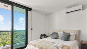 A bedroom with a large bed, a view of greenery through a sliding door, a ceiling fan, and a nightstand with a lamp.