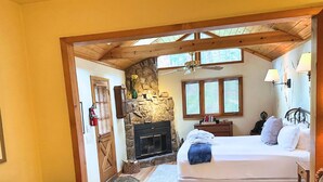 A bedroom with a stone fireplace, a bed with pillows, a nightstand, and a window with blinds. - Chiming Bells Suite at RiverSong Inn in Estes Park - Riverside in Rocky Mountain National Park (Estes Park)